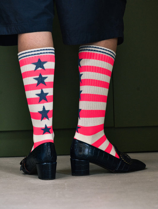 A person's legs wearing vibrant pink and white striped knee-high socks with navy blue stars, paired with black croc-embossed loafers and dark navy shorts, set against a dark green background.