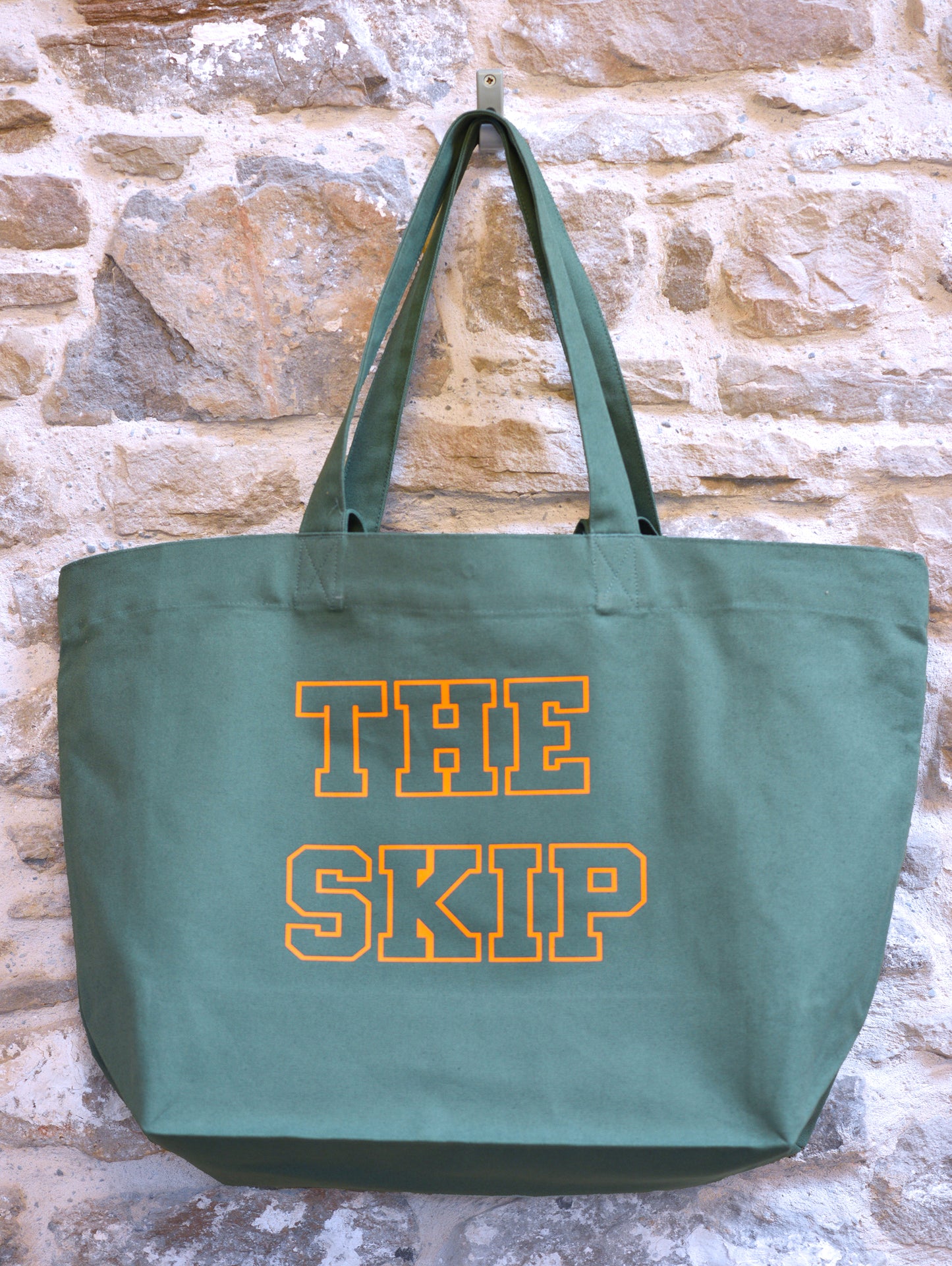 Green tote bag with 'THE SKIP' text on a stone wall background