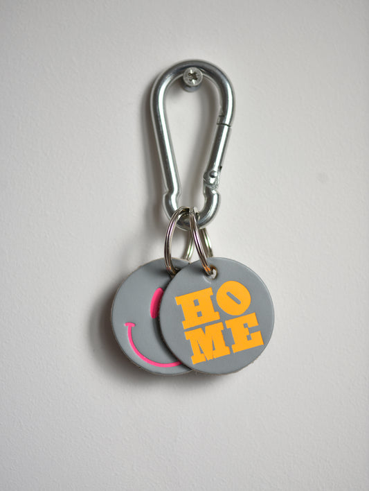 Keychain with 'HOME' and smiley face design on gray tags with a carabiner.