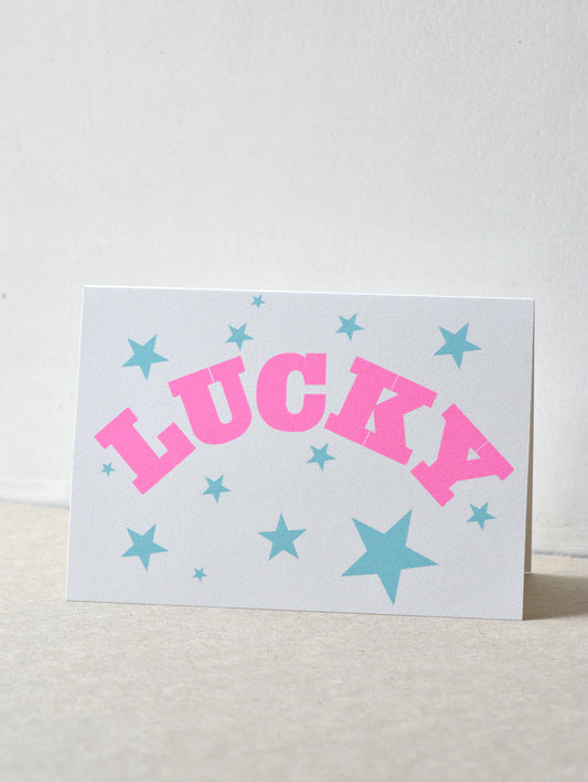Card with 'LUCKY' in pink and stars in blue on a light gray background