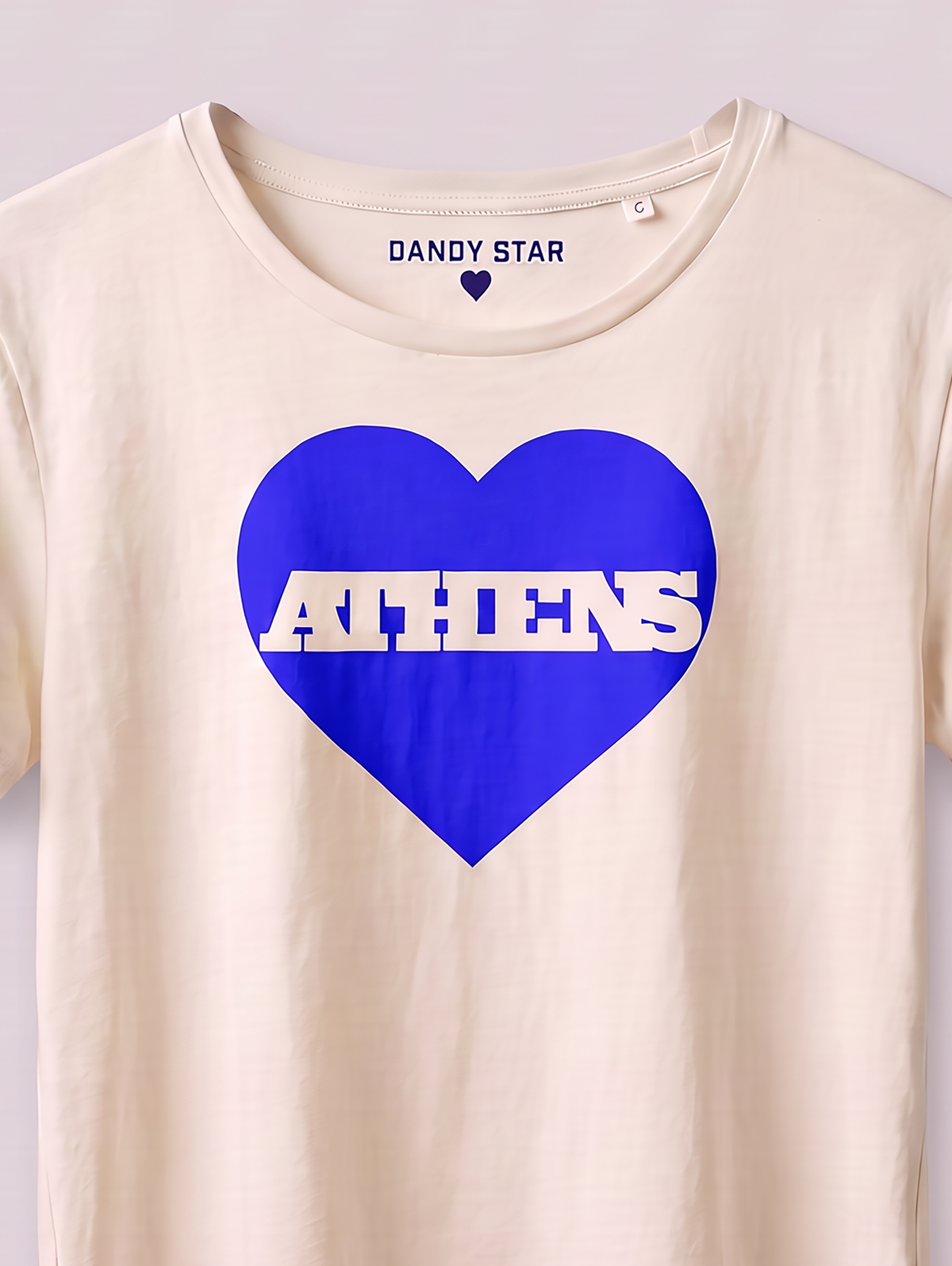 A cream-colored short-sleeve t-shirt featuring a large blue heart graphic on the front with the word 'ATHENS' printed in white across it. The inside collar shows the brand name 'DANDY STAR'.