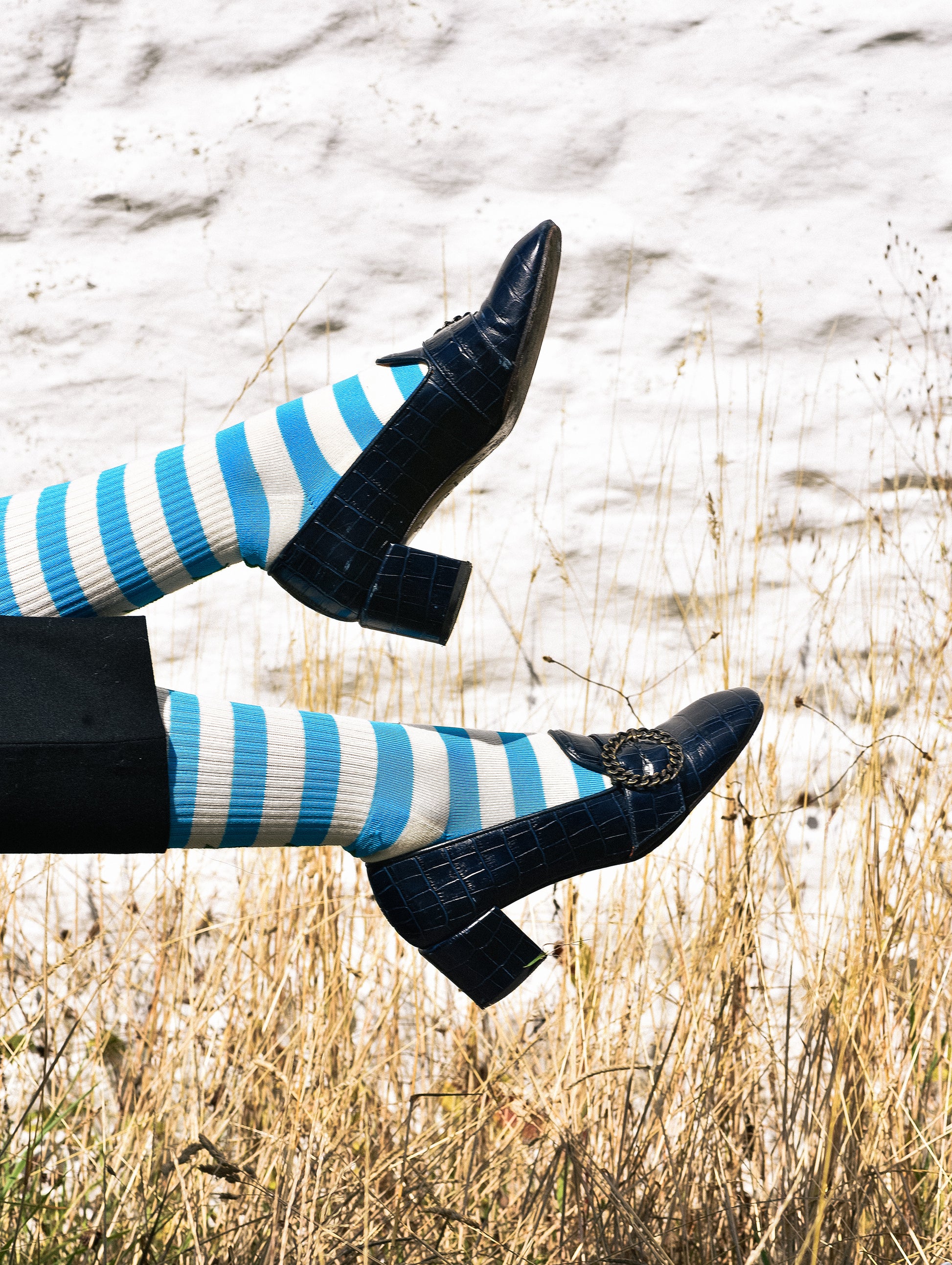 A person's legs kicked up in the air outdoors, wearing blue and white striped tights and dark navy block-heel mules with a crocodile-embossed texture.