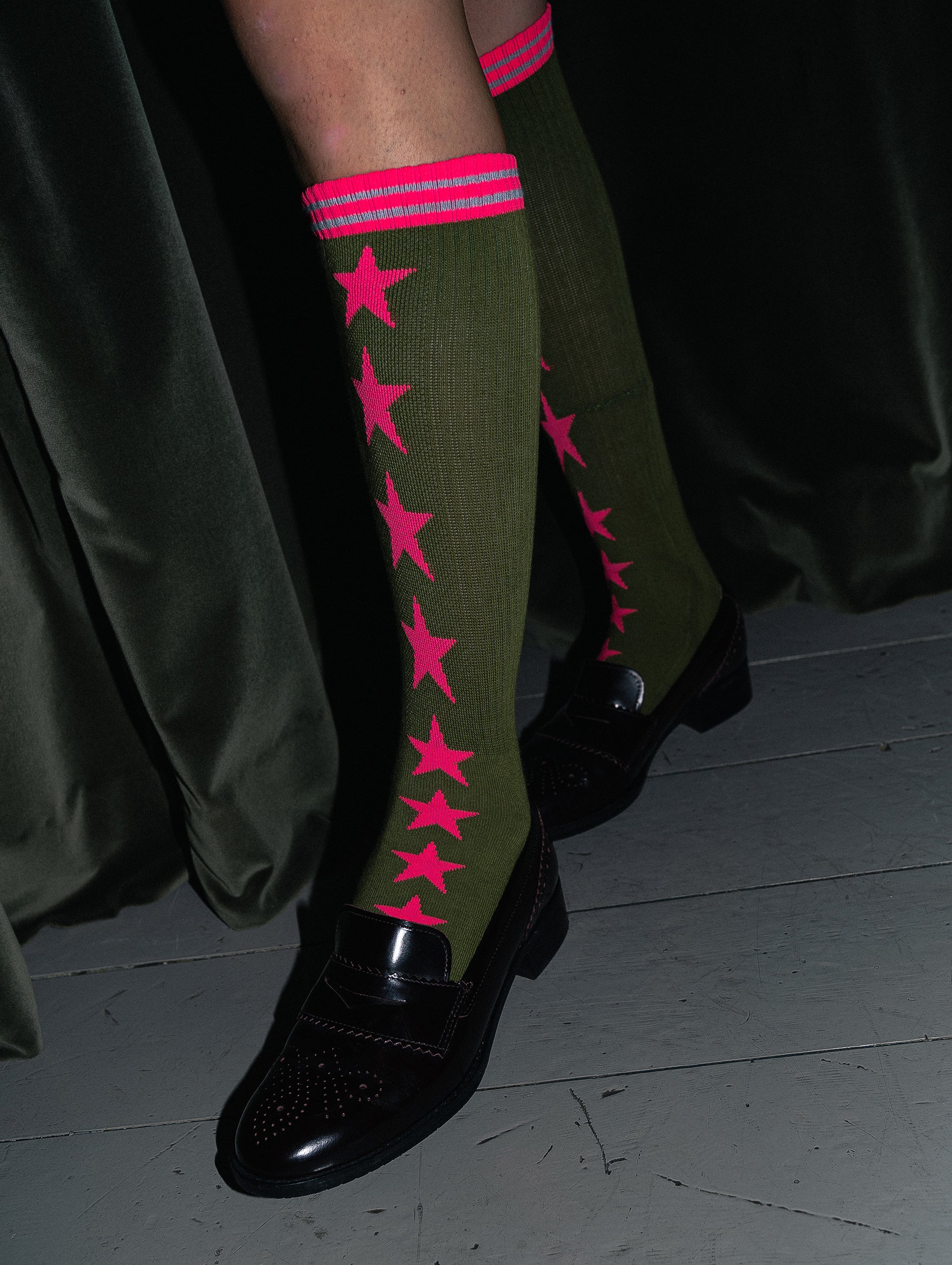Green knee-high socks with pink star patterns worn by a person on a dark background