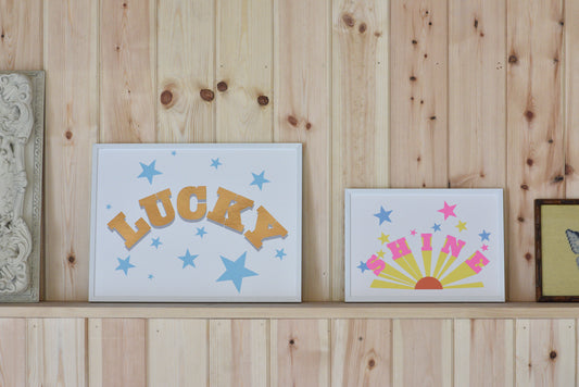 Two framed artworks on a wooden shelf with 'LUCKY' text and star designs.