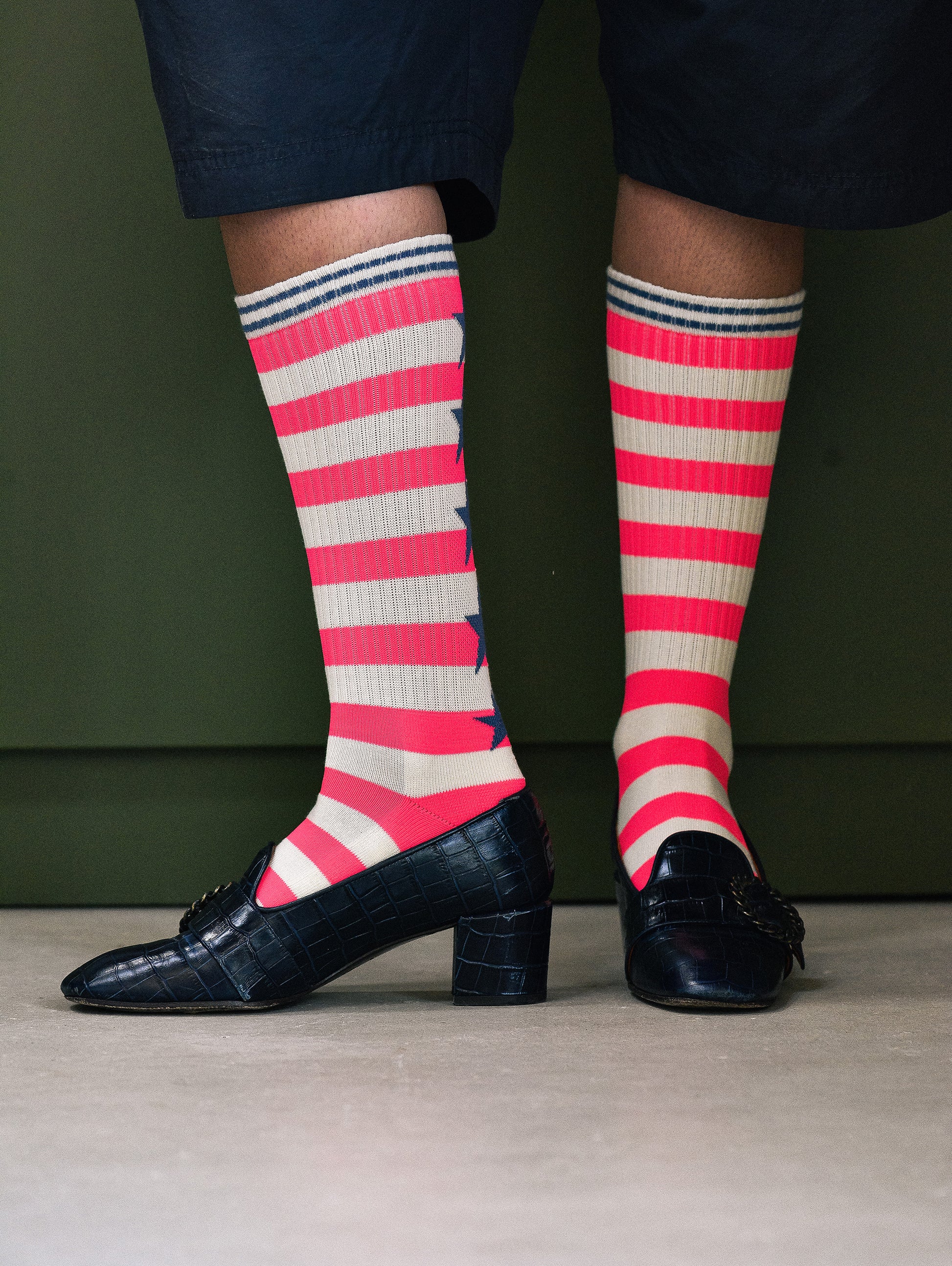 A person wearing bright pink and off-white striped crew socks with navy blue stars, paired with black block-heel loafers and dark navy shorts, standing against a dark olive green background.