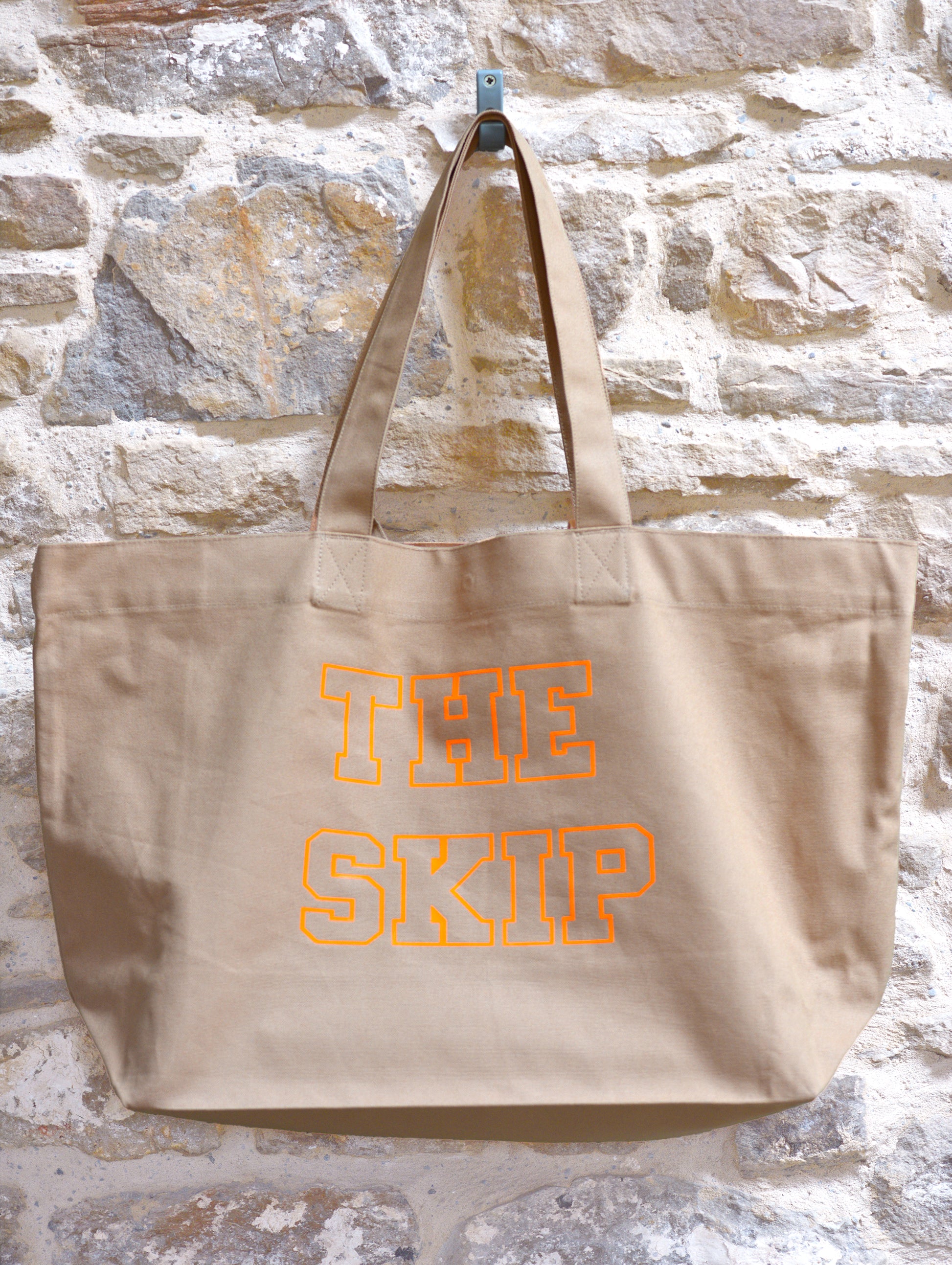 Beige tote bag with 'THE SKIP' text in orange on a stone wall background