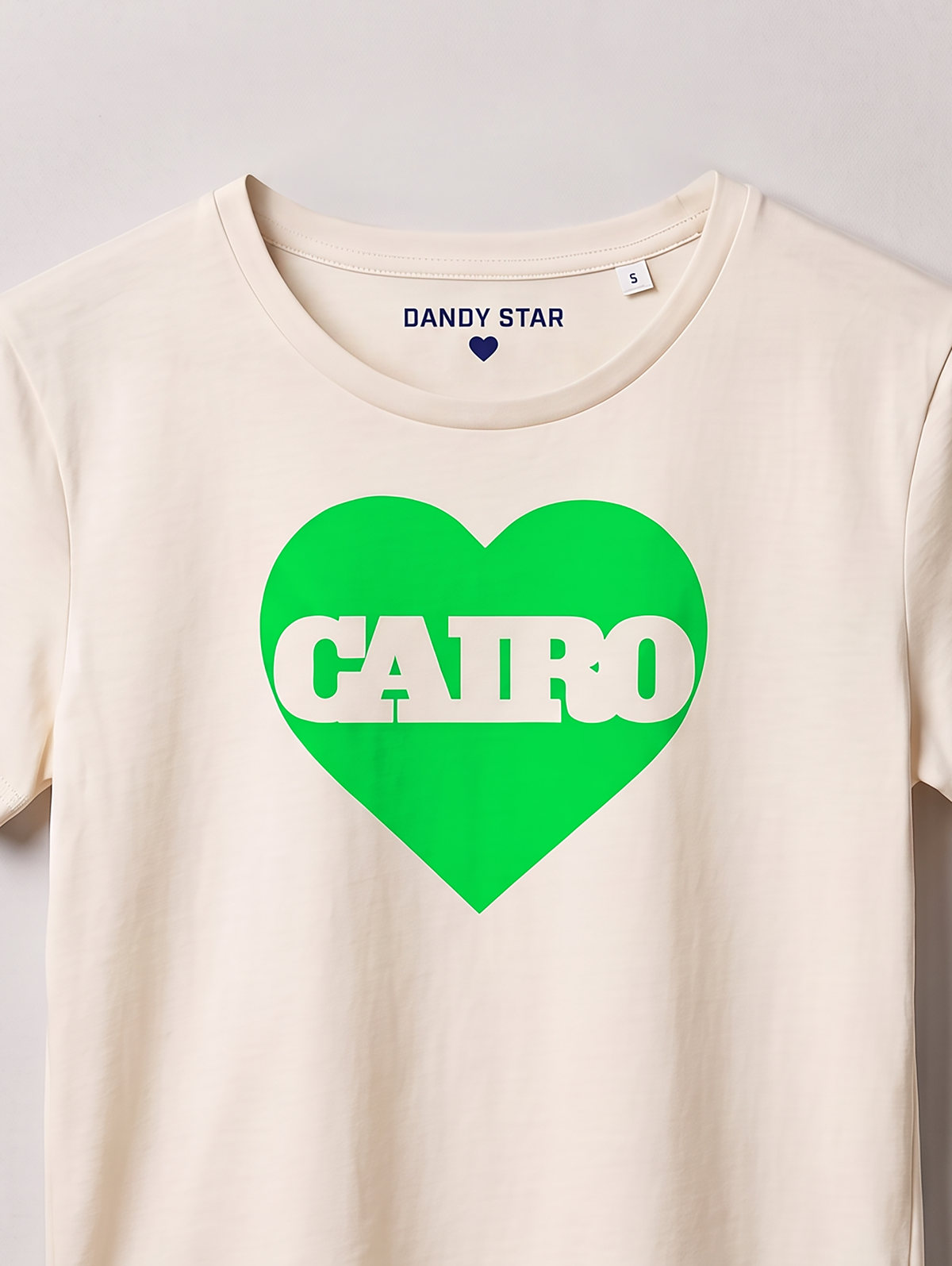 An off-white crewneck t-shirt featuring a bright green heart graphic on the chest with the word 'CAIRO' printed in white across it. The inside collar shows the brand name 'DANDY STAR'.