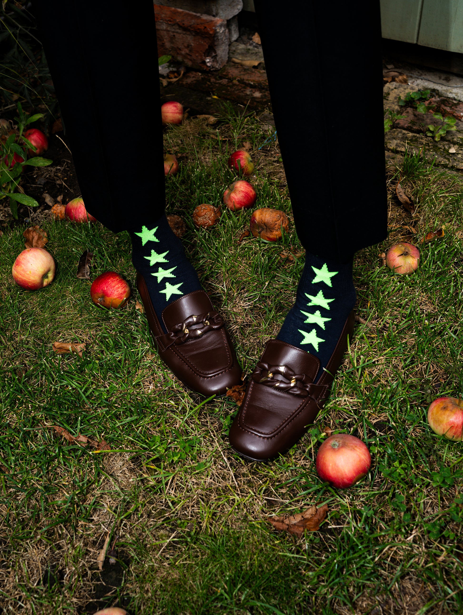 Person wearing brown loafers with green star socks on a grassy ground with apples.